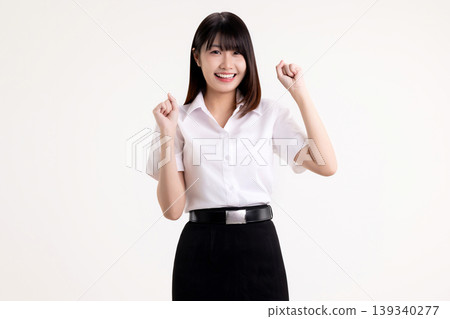 Joyful young Asian woman in student uniform raising clenched fists in victory gesture. Happy female university student celebrating success, achievement, or winning, isolated on white background. 139340277