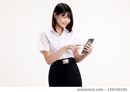 Happy young Asian woman in student uniform using smartphone and touching the screen. Smiling female university student holding mobile phone and browsing social media, isolated on white background. 139340280