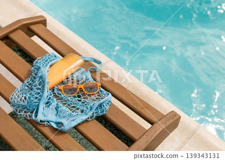 Summer essentials: sunscreen sunglasses and net bag on a wooden chair by a blue swimming pool 139343131
