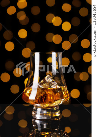 Whiskey in a tulip shaped glass with ice cubes, reflecting golden light against a dark background with orange bokeh, symbolizing a celebratory moment, tasting, or sophisticated evening Whiskey in a tulip shaped glass with ice cubes, reflecting golden light against a dark background with orange bokeh, symbolizing a celebratory moment, tasting, or sophisticated evening 139343654
