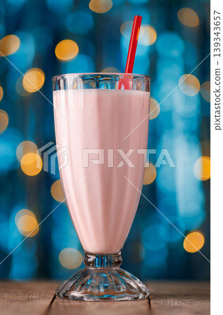 Strawberry milkshake with red straw in tall glass, serving as cool refreshing dessert beverage, sitting on wooden table with sparkling bokeh lights 139343657