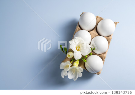 Easter eggs and flowers. White eggs in cardboard box and Freesia white spring flower, flat lay on blue background with copy space Easter eggs and flowers. White eggs in cardboard box and Freesia white spring flower, flat lay on blue background with copy space 139343756
