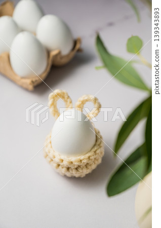 Easter background. Green eucalyptus, white egg in a knitted bunny egg holder. Spring time. Minimalistic Easter background. Green eucalyptus, white egg in a knitted bunny egg holder. Spring time. Minimalistic 139343893