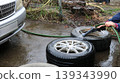 Washing and cleaning car wheels with a water jet from a hose with a watering can attachment, lying on the asphalt next to the car, caring for wheels when changing tires 139343990
