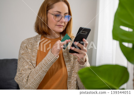 Woman holding smart phone and credit card at home, sitting on the sofa Woman holding smart phone and credit card at home, sitting on the sofa 139344051