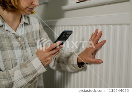 Woman holding smartphone next to heating thermostat, increasing heating costs 139344053