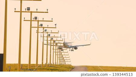 Passenger airplane approaching runway aligned with airport approach lighting system during golden hour. Perspective view of aviation infrastructure. 139344513