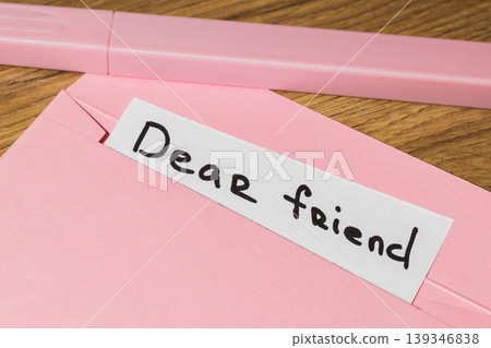 Pink envelope with a white note that reads dear friend on a wooden table with a pink pen Pink envelope with a white note that reads dear friend on a wooden table with a pink pen 139346838