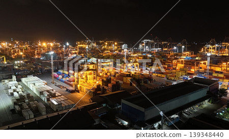 High-angle aerial view of a busy commercial container port terminal at night, featuring colorful shipping crates stacked under bright industrial lights with cranes operating in the background. 139348440