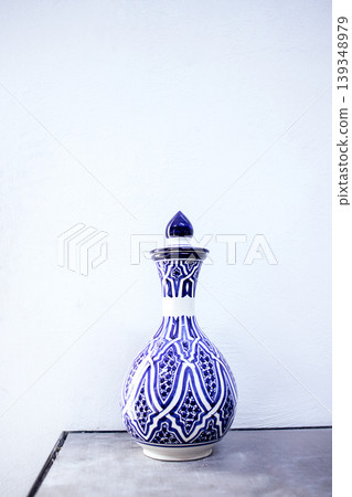 Decorative ceramic vase with intricate blue and white patterns, featuring a narrow neck and a pointed lid, placed on a metallic surface against a plain white background Decorative ceramic vase with intricate blue and white patterns, featuring a narrow neck and a pointed lid, placed on a metallic surface against a plain white background 139348979