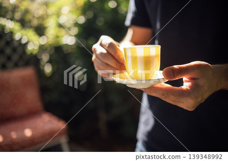 Person holding a yellow striped cup and saucer in a garden setting with blurred greenery and a patterned chair visible in the background Person holding a yellow striped cup and saucer in a garden setting with blurred greenery and a patterned chair visible in the background 139348992