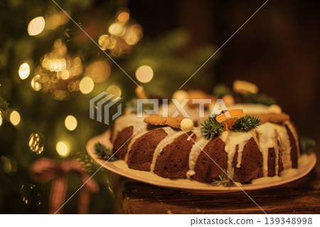 Festive bundt cake decorated with icing, orange slices, and pine branches on a wooden table, with a Christmas tree and warm bokeh lights in the background 139348998