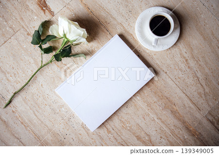 White notebook with blank pages placed beside a cup of black coffee and a single white rose on a textured stone surface, creating a serene and inviting workspace atmosphere White notebook with blank pages placed beside a cup of black coffee and a single white rose on a textured stone surface, creating a serene and inviting workspace atmosphere 139349005