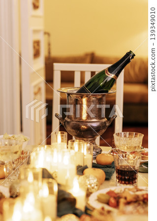 Elegant dining table set for celebration, featuring champagne in silver ice bucket, surrounded by candles, desserts, and festive decor, creating a warm and inviting atmosphere Elegant dining table set for celebration, featuring champagne in silver ice bucket, surrounded by candles, desserts, and festive decor, creating a warm and inviting atmosphere 139349010