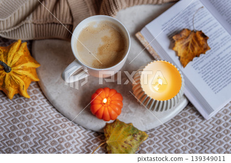 Warm coffee cup with frothy surface, surrounded by autumn leaves, a lit candle, and a cozy book, creating a serene atmosphere for fall relaxation and comfort Warm coffee cup with frothy surface, surrounded by autumn leaves, a lit candle, and a cozy book, creating a serene atmosphere for fall relaxation and comfort 139349011