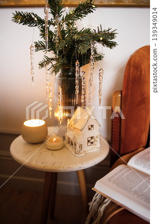 Festive holiday decoration featuring a miniature house, candle, and evergreen arrangement on a cozy table, creating a warm and inviting atmosphere for seasonal celebrations Festive holiday decoration featuring a miniature house, candle, and evergreen arrangement on a cozy table, creating a warm and inviting atmosphere for seasonal celebrations 139349014
