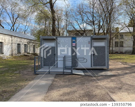 Public toilet facility with accessible entrance and WC signage stands in park area with trees and buildings. Urban infrastructure, public service, accessibility, city environment, outdoor facility 139350796