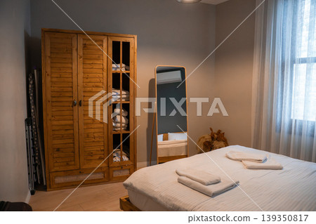 Apartment bedroom interior with wooden wardrobe, standing mirror, bed with linens and folded towels in modern space. Interior design, hospitality concept, real estate, home lifestyle, accommodation 139350817