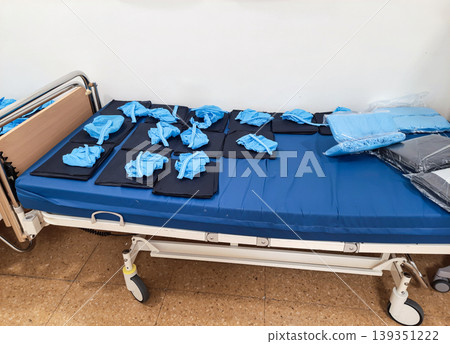 A hospital bed is set with multiple sets of sterile blue surgical scrubs and protective gear. The preparation indicates readiness for medical procedures in a clinical space. A hospital bed is set with multiple sets of sterile blue surgical scrubs and protective gear. The preparation indicates readiness for medical procedures in a clinical space. 139351222