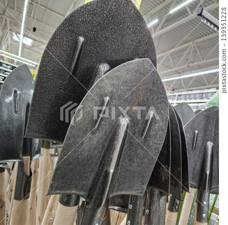Several black metal garden shovels with wooden handles are shown in a hardware store. The shovels are neatly arranged and ready for purchase by customers. Several black metal garden shovels with wooden handles are shown in a hardware store. The shovels are neatly arranged and ready for purchase by customers. 139351228