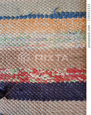 The top view shows a rag rug with striped designs made from various recycled fabric scraps. The rug lies flat against a surface, displaying its unique woven pattern in various colors. The top view shows a rag rug with striped designs made from various recycled fabric scraps. The rug lies flat against a surface, displaying its unique woven pattern in various colors. 139351315