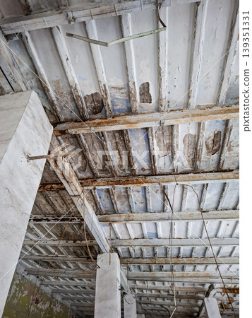 A ceiling with peeling white paint and rusted metal beams in an abandoned interior. The structure displays clear signs of age and neglect. The environment appears desolate. 139351331