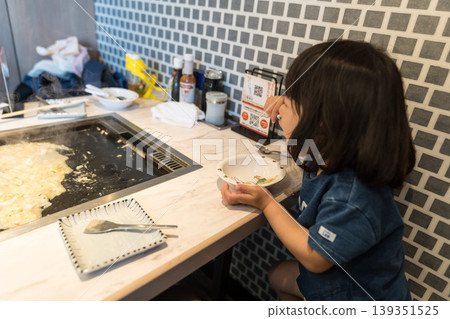 A young Japanese girl eating monjayaki at a teppanyaki restaurant. A young Japanese girl eating monjayaki at a teppanyaki restaurant. 139351525