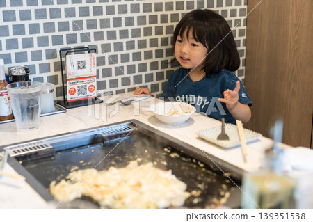 A young Japanese girl eating monjayaki at a teppanyaki restaurant. A young Japanese girl eating monjayaki at a teppanyaki restaurant. 139351538