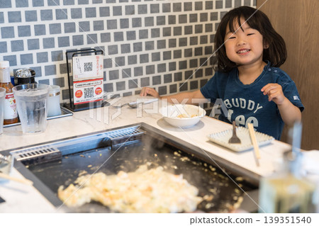 A young Japanese girl eating monjayaki at a teppanyaki restaurant. A young Japanese girl eating monjayaki at a teppanyaki restaurant. 139351540