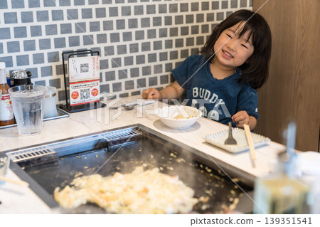 A young Japanese girl eating monjayaki at a teppanyaki restaurant. A young Japanese girl eating monjayaki at a teppanyaki restaurant. 139351541