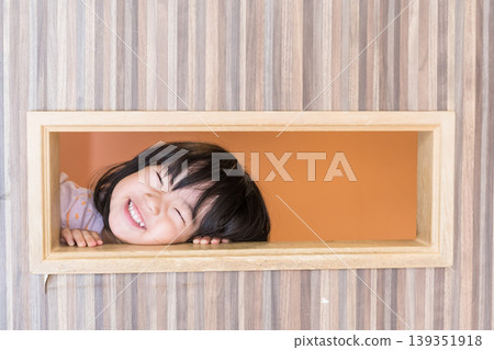 A Japanese toddler girl peeks out from a hole in the wooden frame of a kids' room with a big smile on her face. A Japanese toddler girl peeks out from a hole in the wooden frame of a kids' room with a big smile on her face. 139351918