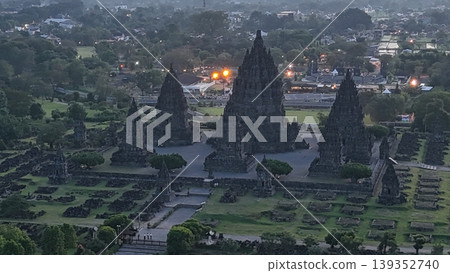 An enchanting evening aerial shot of the Prambanan Hindu Temple complex in Indonesia, featuring its iconic towering stone spires illuminated against a misty twilight sky and a sprawling cityscape. An enchanting evening aerial shot of the Prambanan Hindu Temple complex in Indonesia, featuring its iconic towering stone spires illuminated against a misty twilight sky and a sprawling cityscape. 139352740