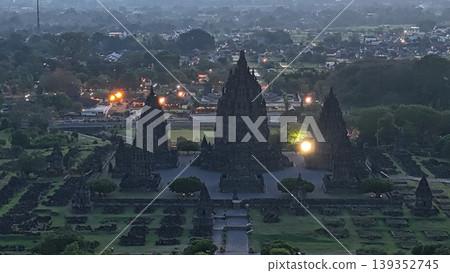 An enchanting evening aerial shot of the Prambanan Hindu Temple complex in Indonesia, featuring its iconic towering stone spires illuminated against a misty twilight sky and a sprawling cityscape. 139352745