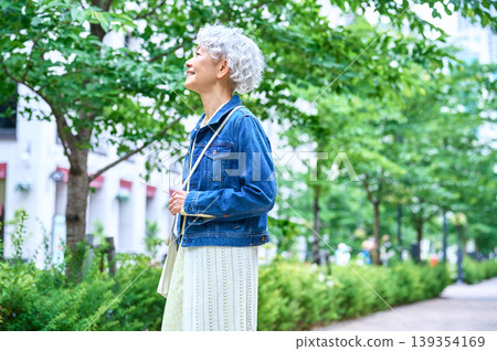 A senior woman looks up at the sky sideways while walking along a tree-lined street. A senior woman looks up at the sky sideways while walking along a tree-lined street. 139354169