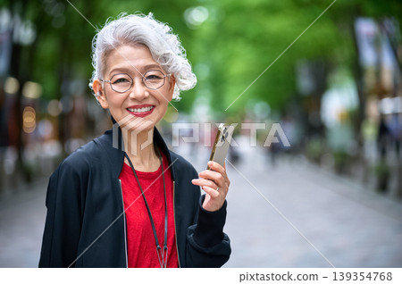 A senior woman with a smartphone and a smile walks down a tree-lined street. A senior woman with a smartphone and a smile walks down a tree-lined street. 139354768
