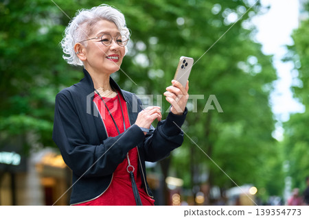 一位老婦人在綠樹成蔭的街道上使用智慧型手機 一位老婦人在綠樹成蔭的街道上使用智慧型手機 139354773