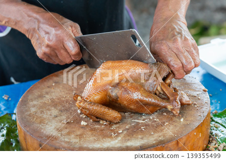 Hands slicing roasted duck on wooden chopping board with cleaver showing traditional cooking process rich texture and street food preparation Hands slicing roasted duck on wooden chopping board with cleaver showing traditional cooking process rich texture and street food preparation 139355499