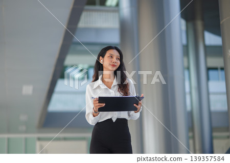 Confident businesswoman holds a clipboard and pen while looking away in a modern office atrium, ideal for corporate strategy, project planning, leadership, and business career growth. Confident businesswoman holds a clipboard and pen while looking away in a modern office atrium, ideal for corporate strategy, project planning, leadership, and business career growth. 139357584