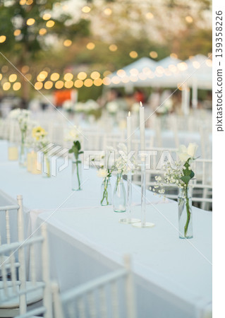 Elegant wedding long table with candle, white rose flower and white chairs in the garden Elegant wedding long table with candle, white rose flower and white chairs in the garden 139358226