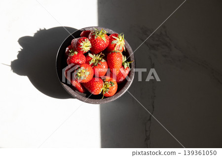 Bowl of fresh strawberries on white marble surface with soft shadows. Top view of ripe red fruit in black bowl. Healthy eating and summer freshness concept for nutritious recipes and diet inspiration. Bowl of fresh strawberries on white marble surface with soft shadows. Top view of ripe red fruit in black bowl. Healthy eating and summer freshness concept for nutritious recipes and diet inspiration. 139361050