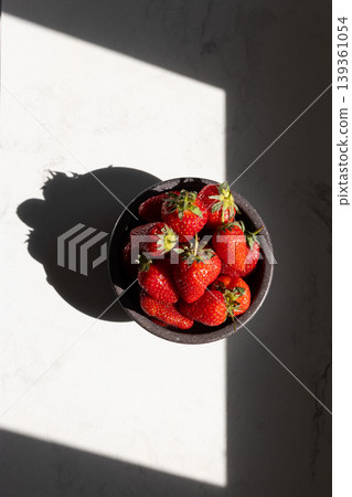 Fresh red strawberries in black bowl on white marble surface. Sunlight casting dramatic shadows. Healthy summer fruit and natural food concept for recipes and nutrition. Fresh red strawberries in black bowl on white marble surface. Sunlight casting dramatic shadows. Healthy summer fruit and natural food concept for recipes and nutrition. 139361054