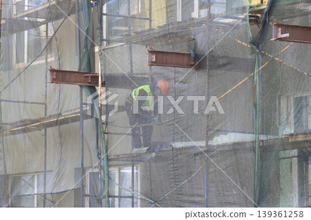 Construction worker in safety vest and hard hat working on high-rise scaffolding behind a protective net Construction worker in safety vest and hard hat working on high-rise scaffolding behind a protective net 139361258