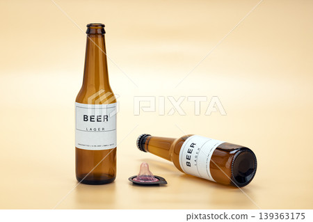 Amber beer lager bottles beside an unrolled pink condom on a warm pastel yellow background, representing safe sex awareness and the risks of alcohol and sexual decision-making 139363175