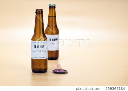 Two brown glass craft lager beer bottles standing beside an unwrapped pink condom on a warm beige studio background, representing safe sex and responsible drinking concept photography Two brown glass craft lager beer bottles standing beside an unwrapped pink condom on a warm beige studio background, representing safe sex and responsible drinking concept photography 139363184