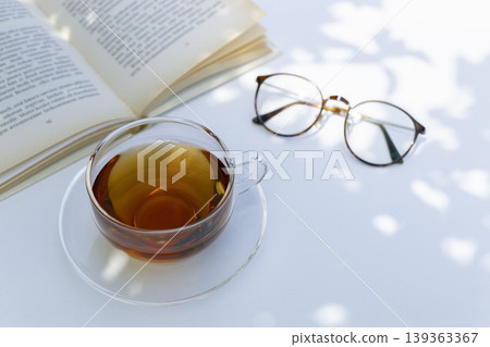 在白色的桌子旁,手捧一杯茶,面前擺著一本打開的書和幾隻杯子,享受一段寧靜的閱讀時光(桌子周圍留有空白區域)。 在白色的桌子旁,手捧一杯茶,面前擺著一本打開的書和幾隻杯子,享受一段寧靜的閱讀時光(桌子周圍留有空白區域)。 139363367