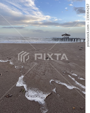 Beach with gentle waves and wooden pier under cloudy sky, serene nature background scene 139364267