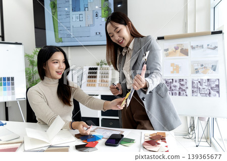 Asian women designers collaborating on interior material selection reviewing architectural plans discussing color palettes for modern home project office 139365677