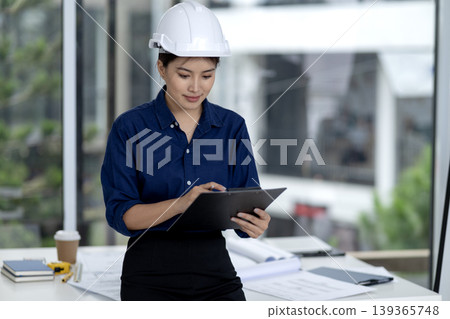 Asian Female Engineer Architect Reviewing Project Plans At Modern Office Wearing Safety Helmet Overseeing Construction Development 139365748