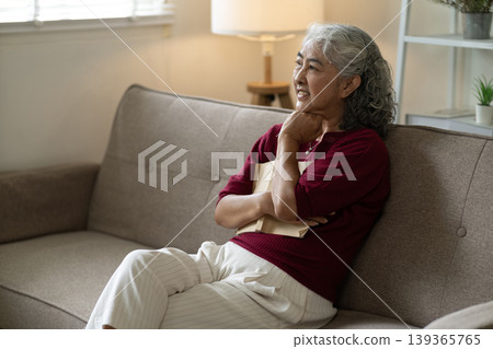 Asian senior woman relaxing reading book at home living room sofa thoughtful expression enjoying quiet leisure moment cozy domestic comfort daily life 139365765
