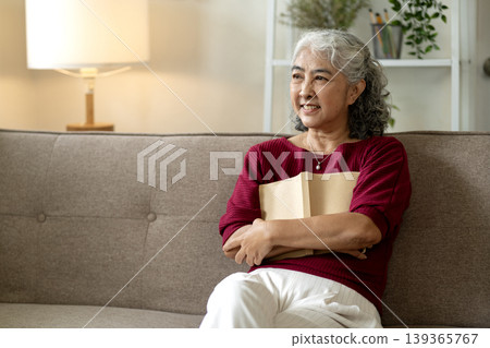 Asian older woman sitting sofa reading book enjoying quiet thoughtful moments at home living room embracing peaceful leisure lifestyle retirement 139365767